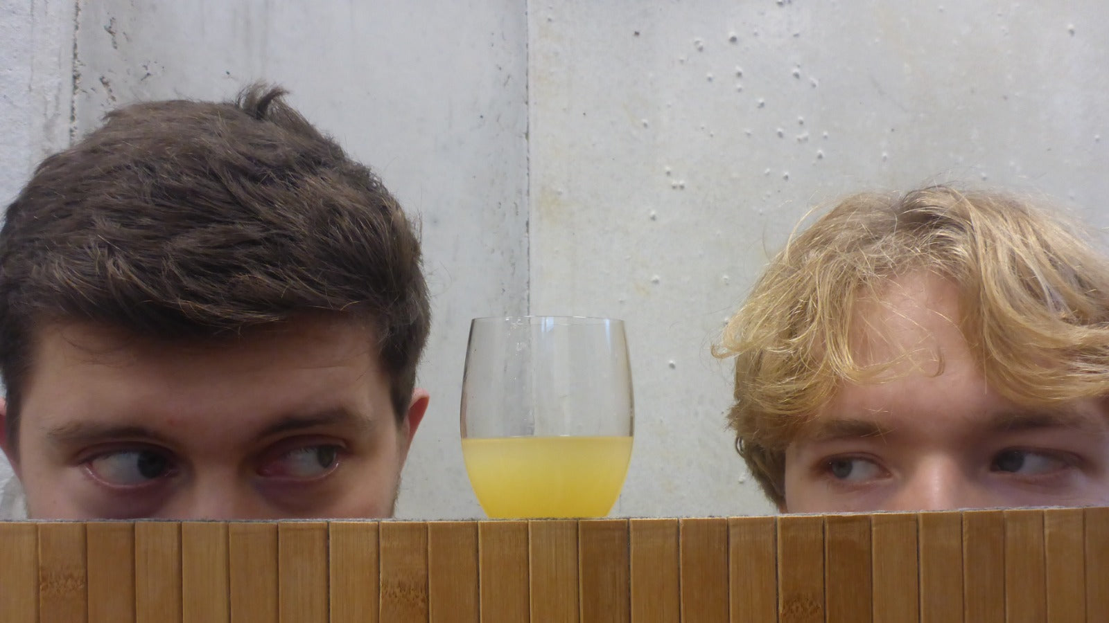 Two people behind a wooden panel with a glass of yellow liquid between them.